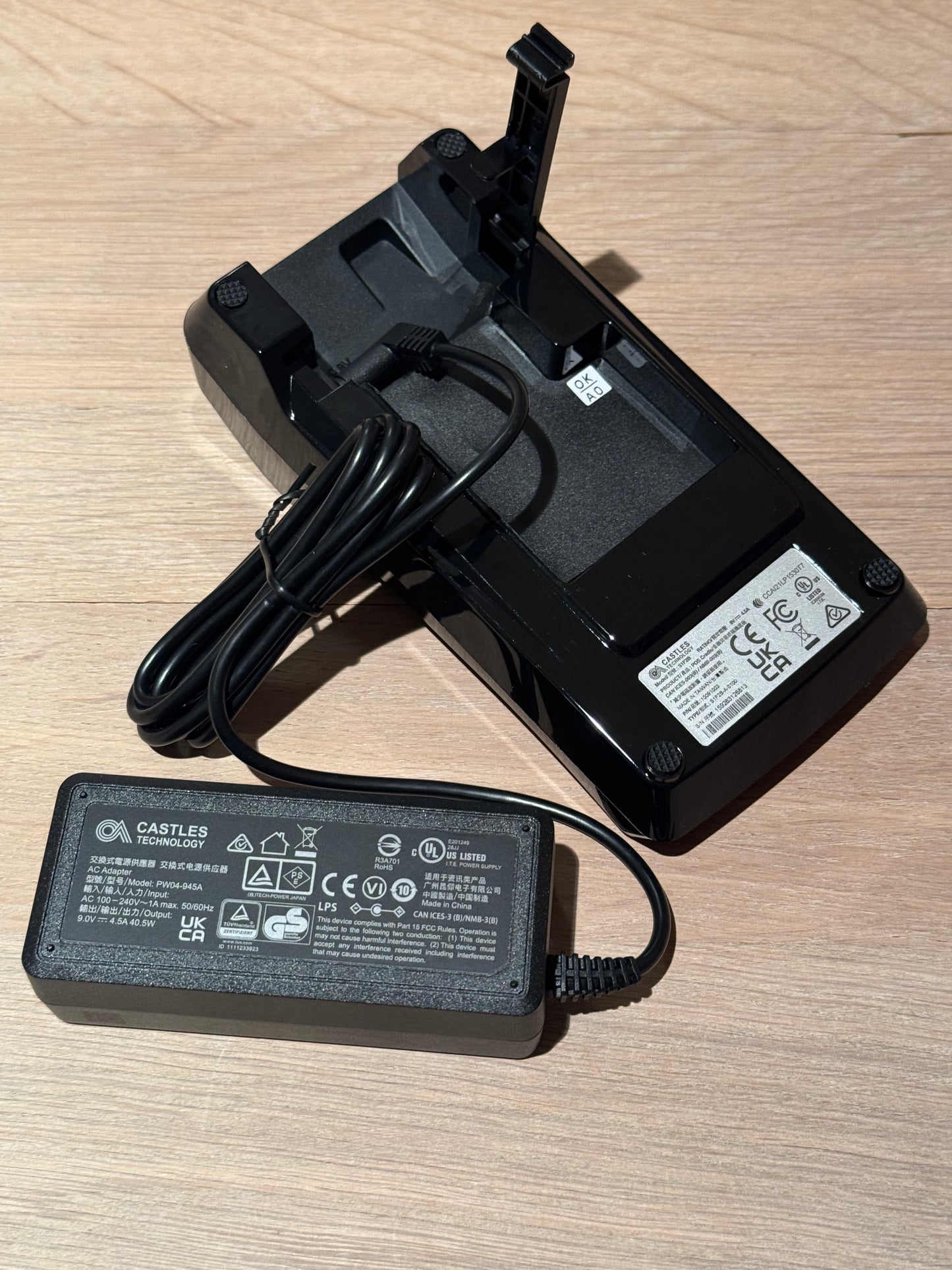 A black Castles Technology battery charger and power adapter. The charger has a built-in battery compartment with a latch, and the separate power adapter is shown in front with its cord and a white specification label.